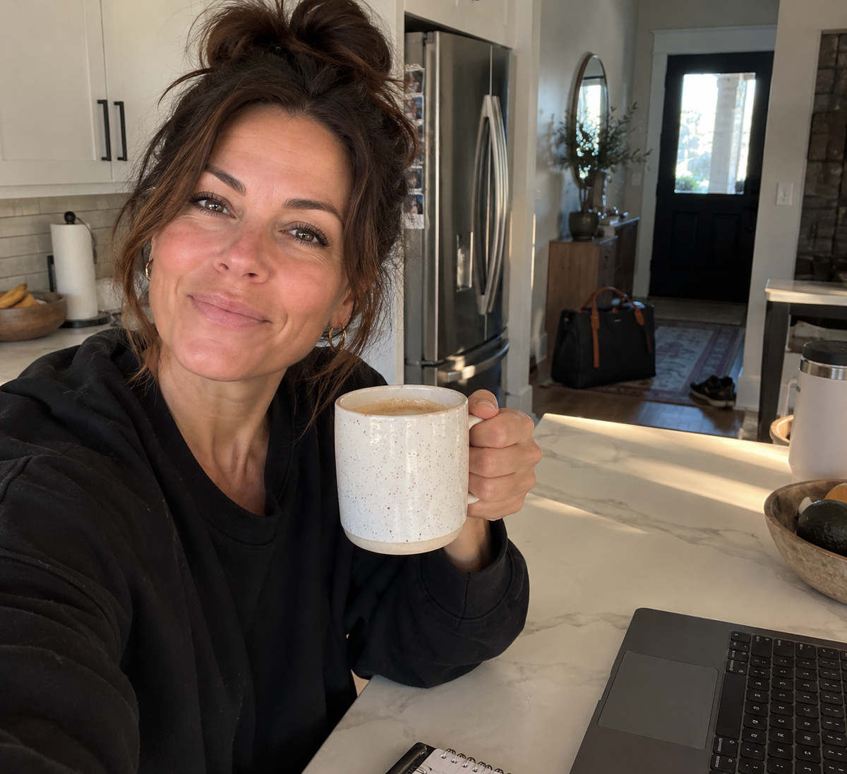 A woman in her forties drinking coffee at a sunlit kitchen counter, looking rested