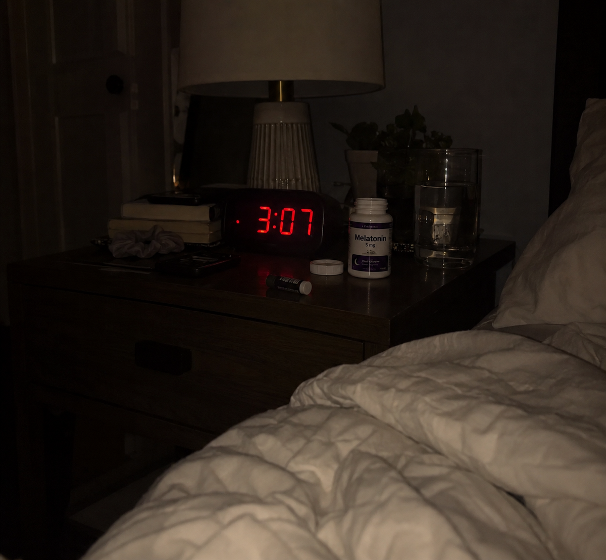 A digital clock glowing 3:07 on a dark nightstand next to a bottle of melatonin gummies